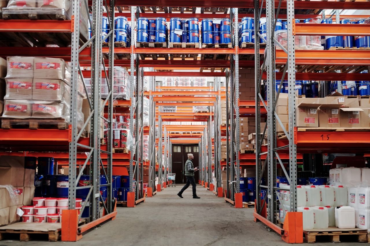 heros-img A man walking through a large industrial warehouse with stacked shelves filled with goods and products.
